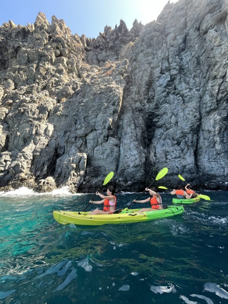 Dos personas en un kayak bajo los acantilados de roca volcánica, sobre agua verde y transparente